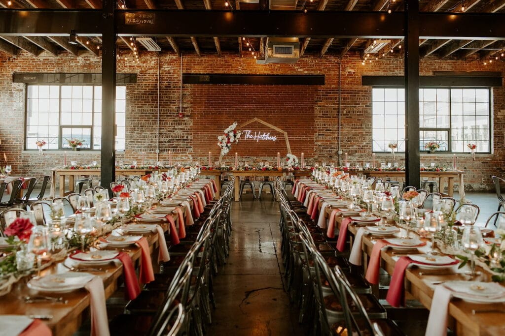 An industrial event space with exposed brick walls and metal beams is set up for a reception with a long wooden guest and head tables decorated in autumn hues