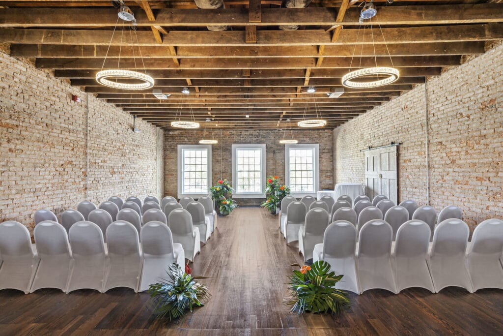 Exposed brick event space with wooden beam ceiling, large bright windows and sliding barn doors set up for wedding ceremony with white covered chairs and tropical flowers at the start and end of the aisle