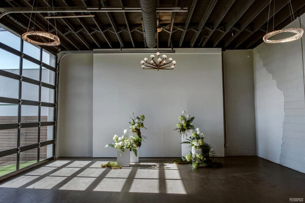 A simple wedding altar of green and white flowers shines in the sunlight from a giant glass garage door in an industrial event space