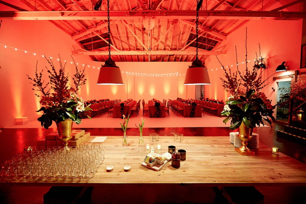 Five long banquet tables draped in red linens and rich floral arrangements in a studio set decorated for a reception with string lights and uplighting against the walls