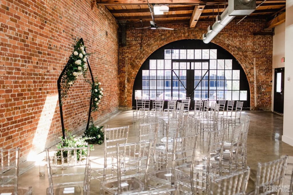 Clear chairs are organized in an industrial venue for a wedding ceremony by a large arched window