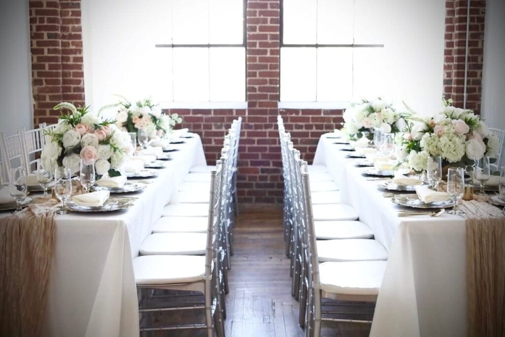 Two long tables draped in white and tan linens and decorated with white, green and pink floral arrangements are lit by a large window in a brick industrial space