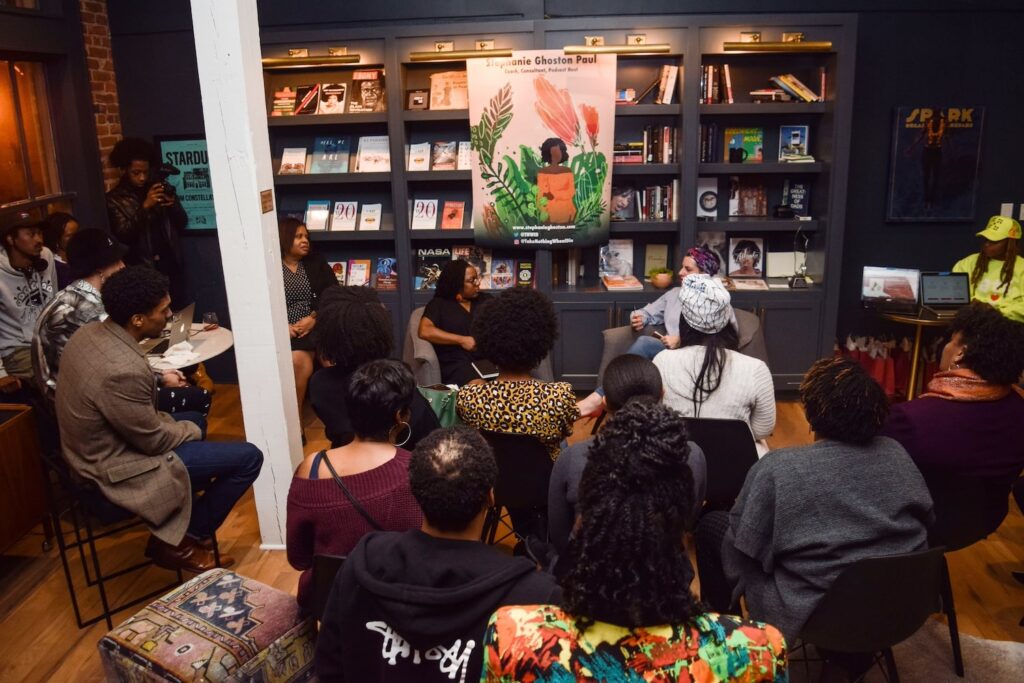 An intimate group gathers for a discussion between two women in a small space with dark-painted bookshelves displaying books and magazines