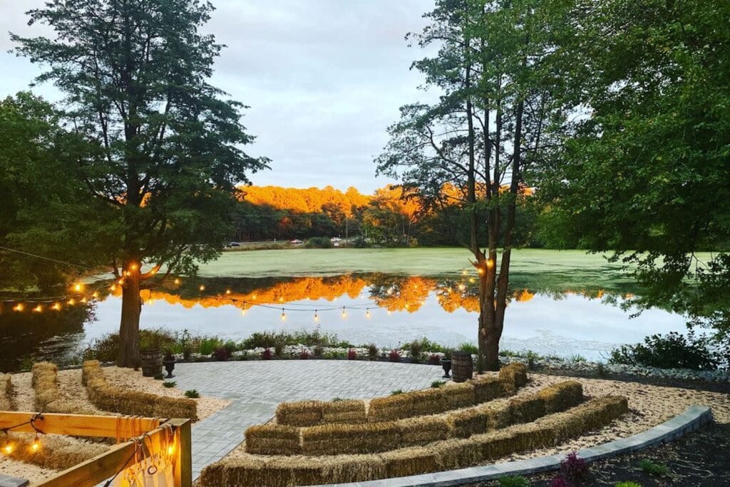 String lights hover above hay bales arranged as seating by a lake for an outdoor wedding ceremony