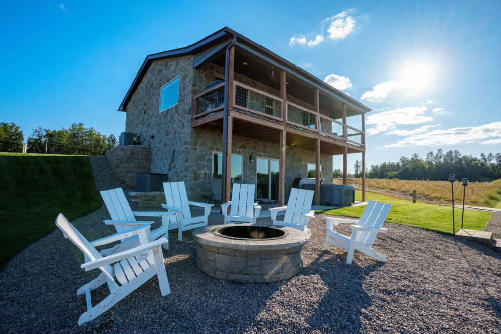A two-story stone lake house sits in the sunshine with a fire pit in the foreground surrounded by white Adirondack chairs