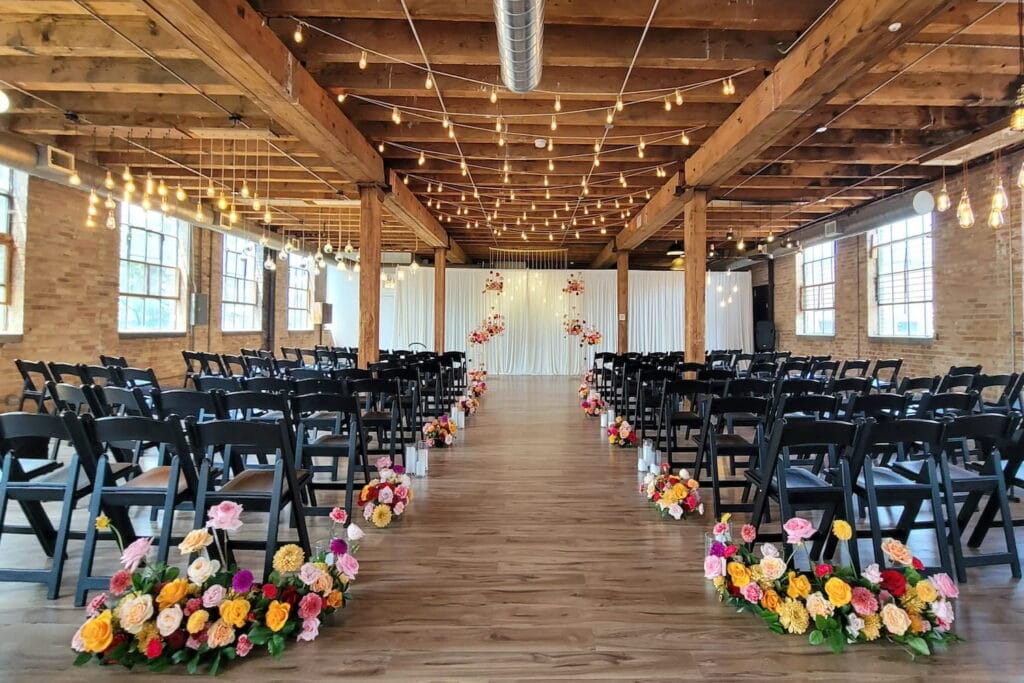 Brightly colored flowers line a ceremony aisle created by two groups of black folding chairs in an industrial loft event space