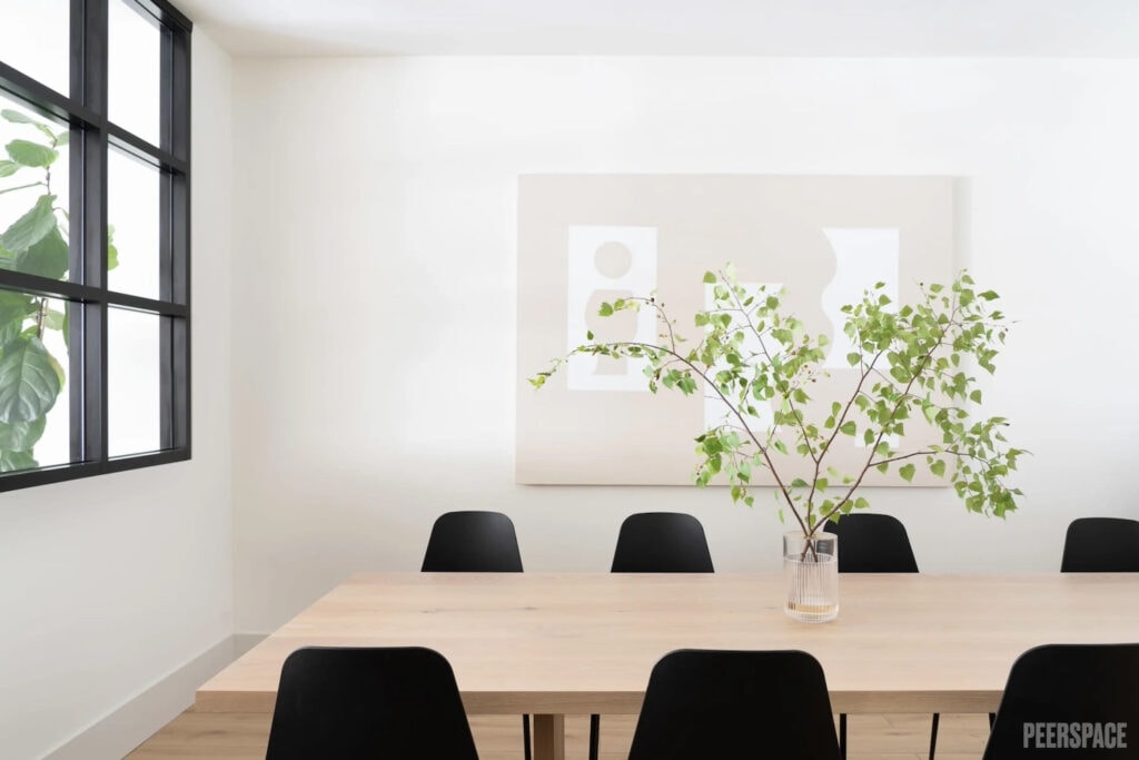 A clear vase with greenery sits in the middle of a light wood conference table in a white room with a black window and black chairs
