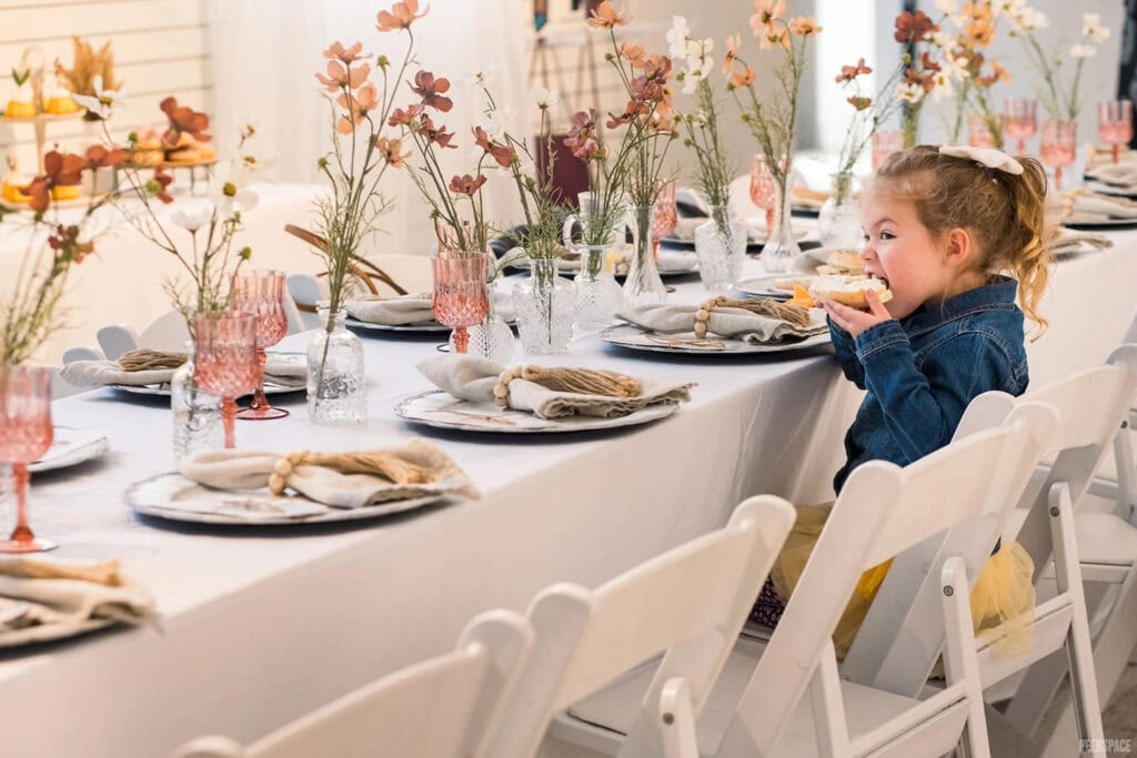 A little girl in a tulle skirt and jean jacket eats a pastry at a long table set for a party