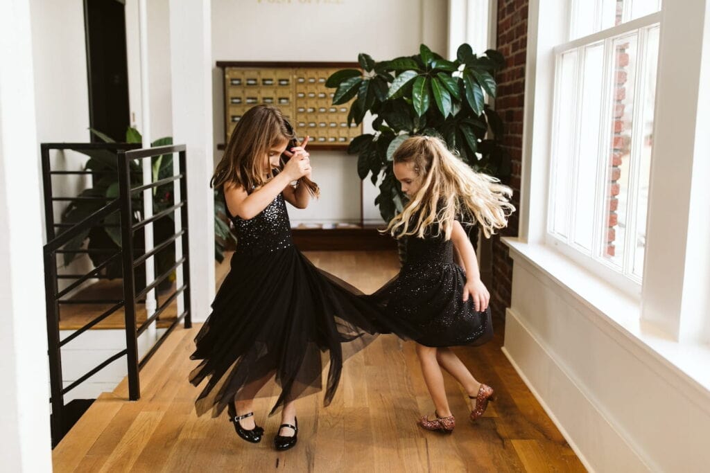 Little girls in black party dresses dance, watching their skirts twirl around them by a bright window