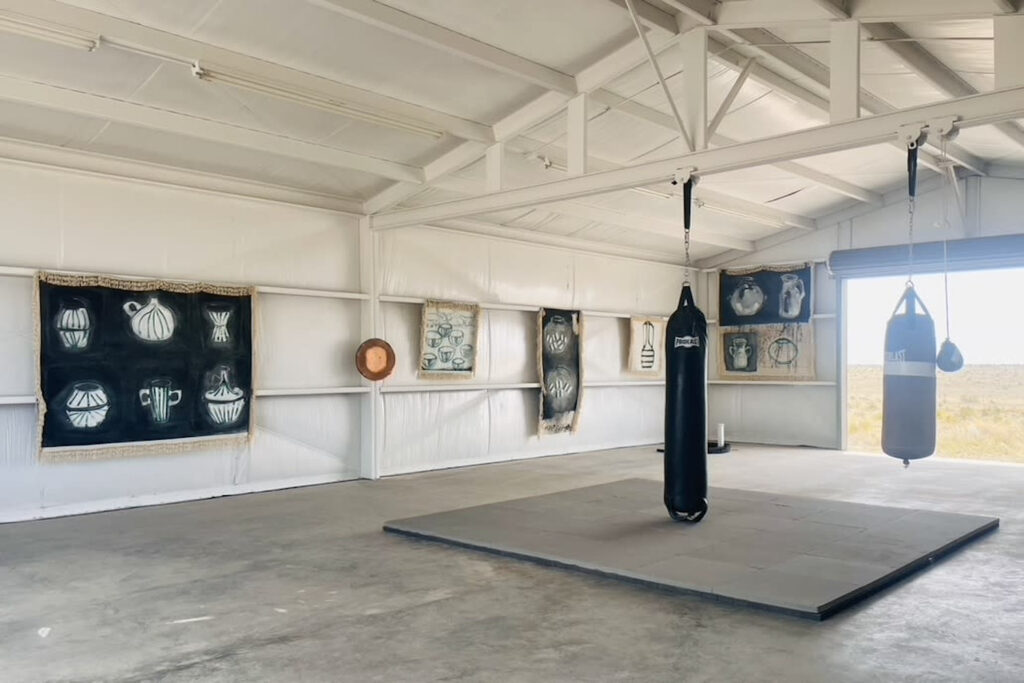 Punching bags hang from beams across an arched ceiling in an all-white garage space with a giant garage door opening up to the outside