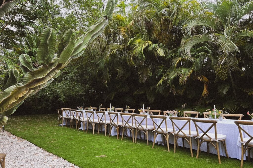 A long banquet table set up outside in white linens with wooden chairs surrounded by palm trees