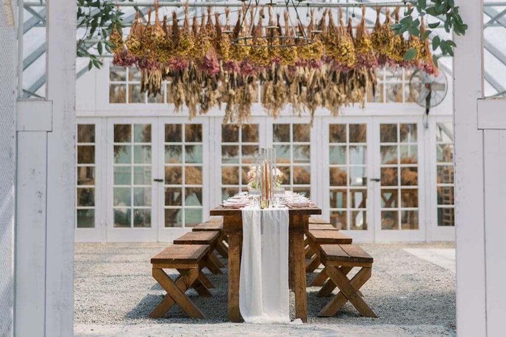 A long wooden table and benches sit below dried flowers in a rustic, brightly lit, open-air venue space