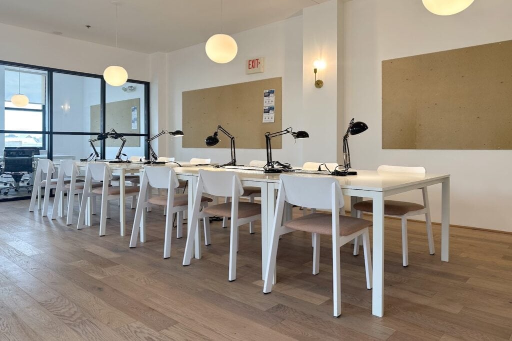 A long white worktable surrounded by white chairs with black table lamps on top sits in a white conference room next to a small office space