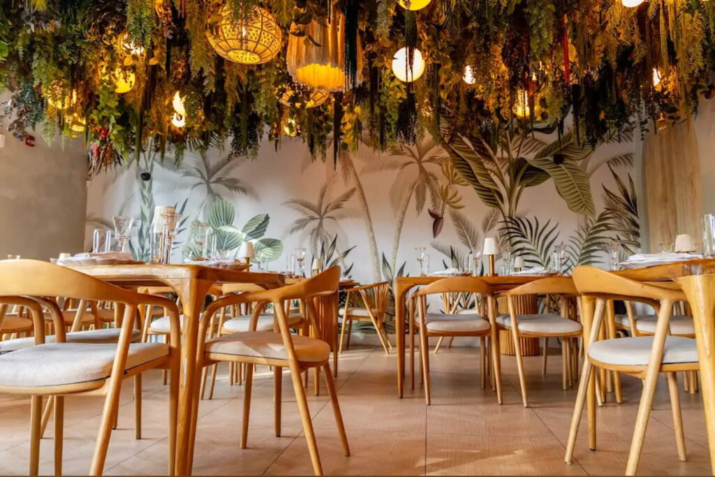 Greenery hangs from the ceiling of a romantic dining space with a mural of palm fronds on one wall and warm wood seating