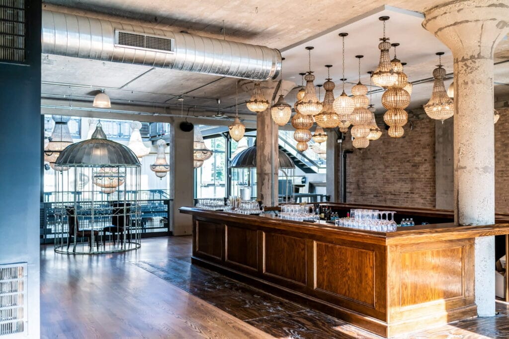 Multiple chandeliers of different shapes and sizes hang from the cement ceiling of an industrial event space with exposed ductwork and brick walls with a warm wooden bar in the center