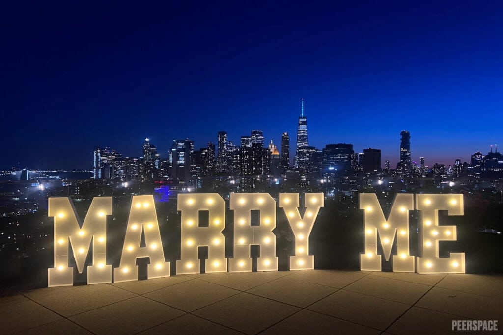 Giant light up letters spelling out Marry Me sit on a rooftop with city views in the background