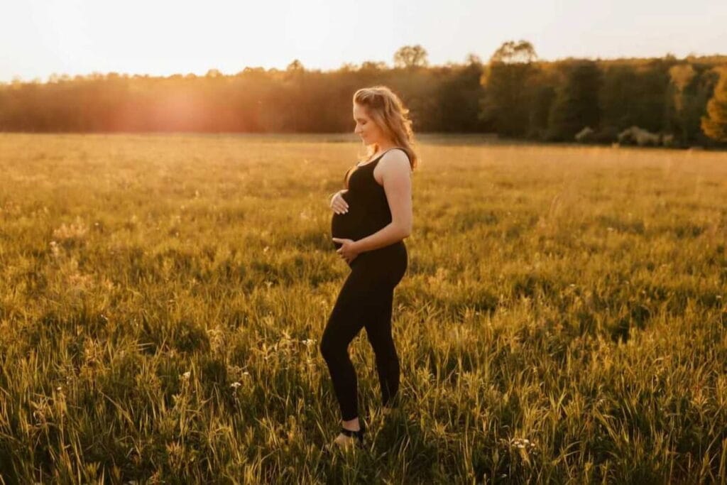 A woman poses in a field cradling her pregnant belly with the sun setting in the distance