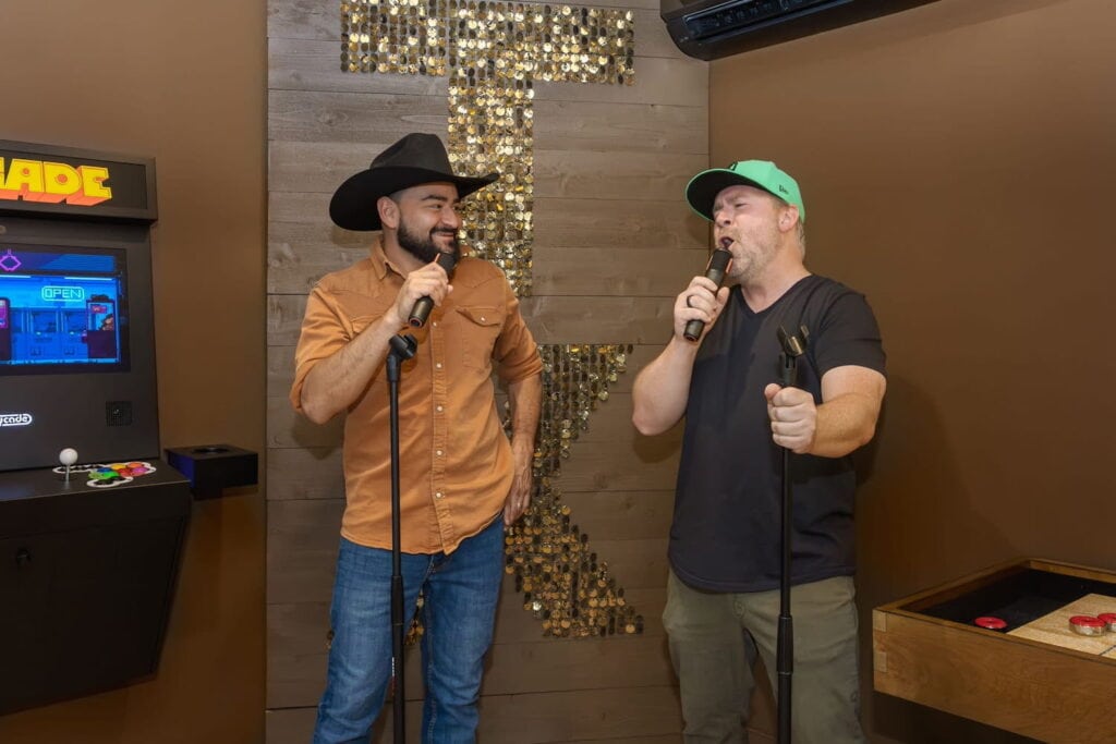 Two men sing at microphones next to an arcade machine