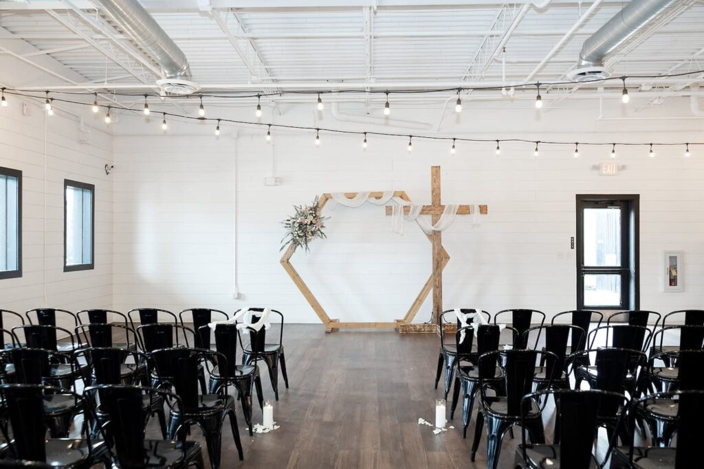 Black metal chairs face an altar decorated with a wooden cross and hexagon draped in flowers and white cloth in an all-white venue with black windows and string lights