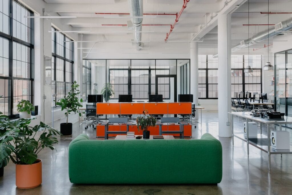 Bright orange storage cabinets punctuate a large work area with multiple seating and desk areas and surrounded by large, paned windows