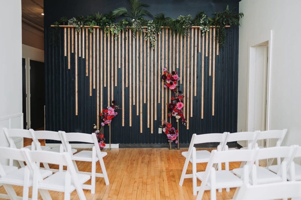 A small, modern wedding ceremony space with white wooden chairs, a black and wood wall and an altar of bright flowers