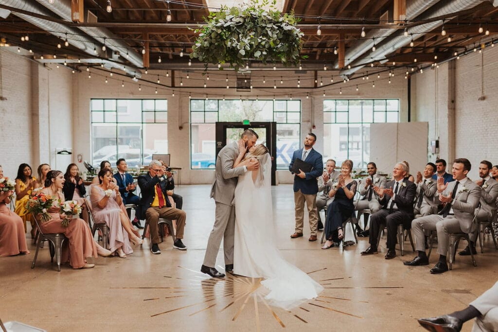 Bride and groom kiss in front of friends and family in a modern industrial venue