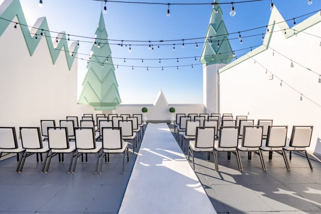 Modern white and black chairs line a simple aisle under string lights on a rooftop punctuated by large tree-like structures adorning the exterior of a building