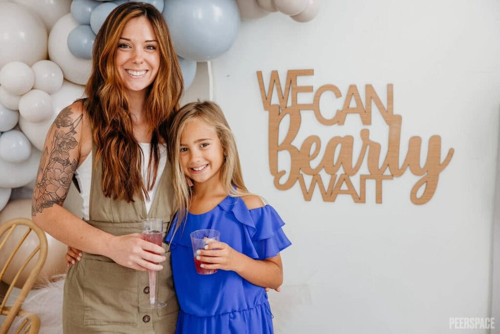 A mom and daughter pose together at a baby event in front of a sign that says We Can Bearly Wait