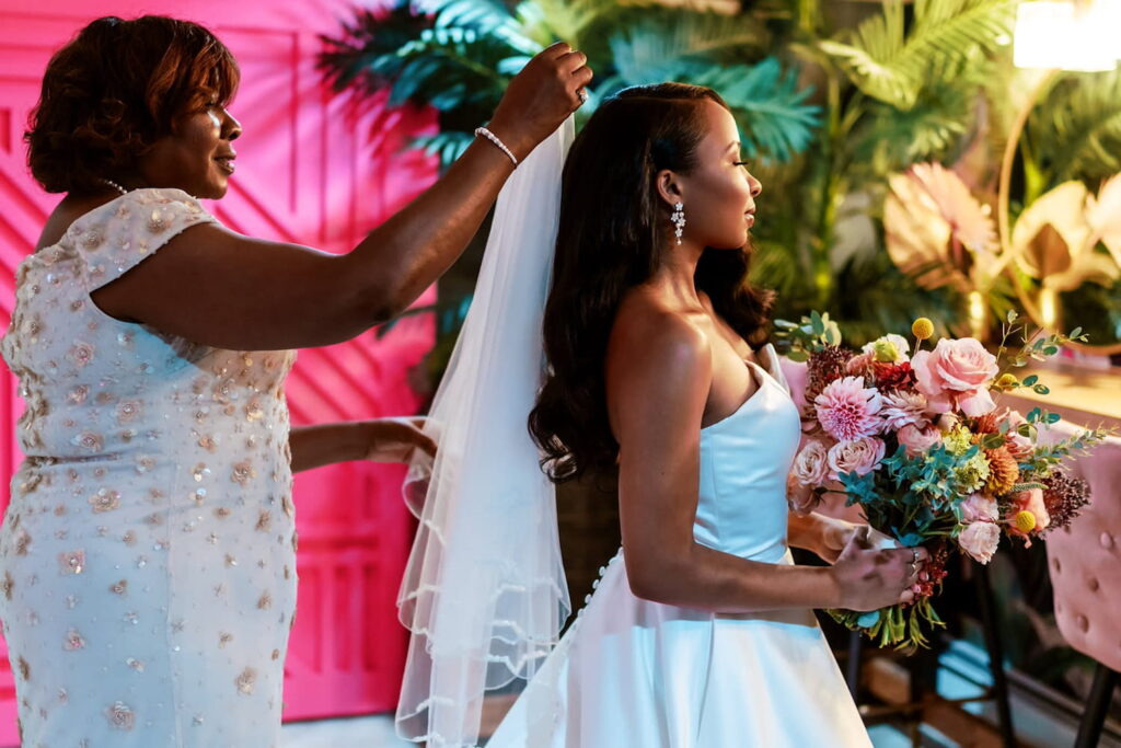 A mother places a veil on a bride holding a large bouquet of pastel-colored flowers in a brightly colored space with a pink wall and large palms