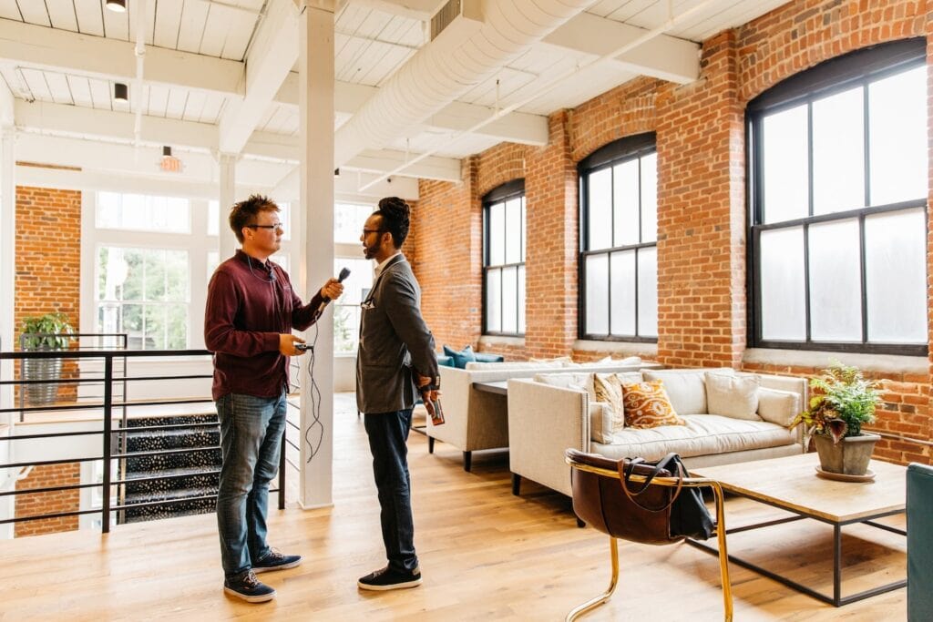 Two individuals conduct an interview in a commercial office space with exposed brick walls and large paned windows.