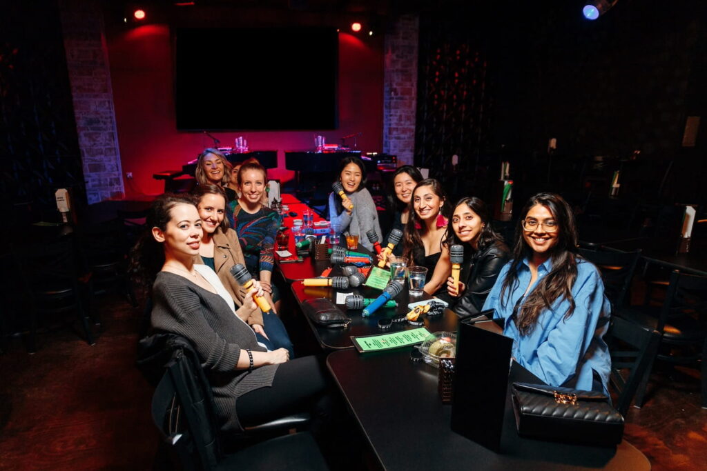 A table of women at music bar smile at the camera holding microphones with pianos on stage in the background