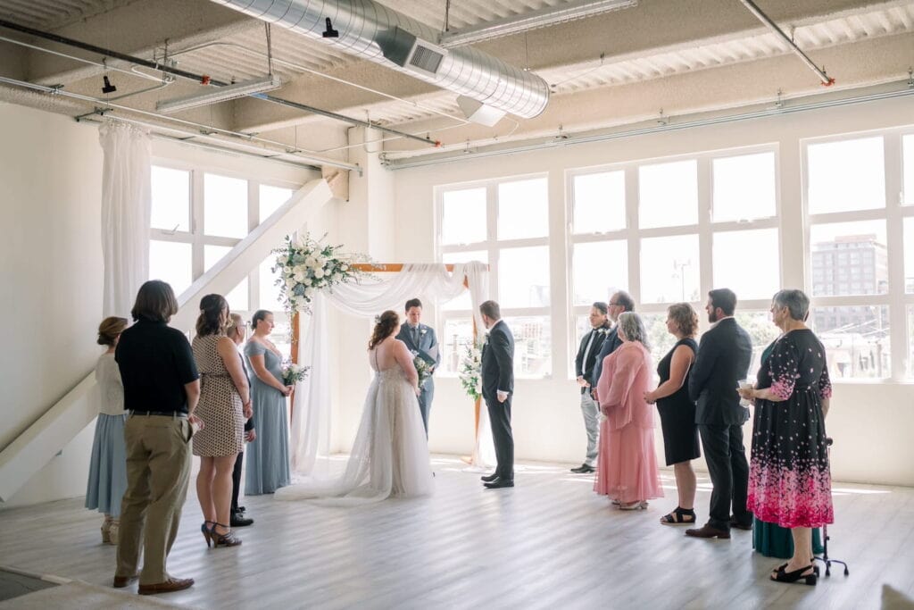 A bride and groom exchange vows in front of friends and family in a bright, airy venue surrounded by windows