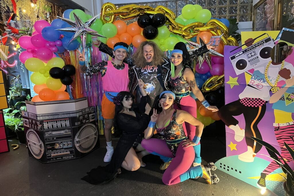 Brightly-dressed crew poses in front of 80s props