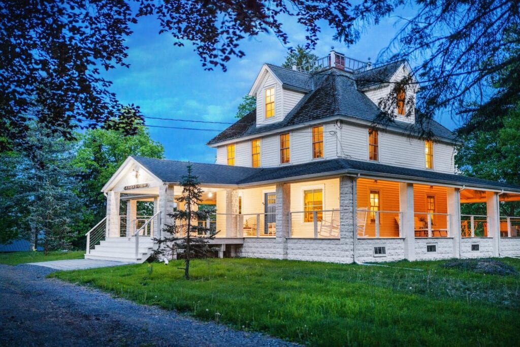 A white farmhouse is lit up a dusk with a large, wraparound porch adorned with rocking chairs