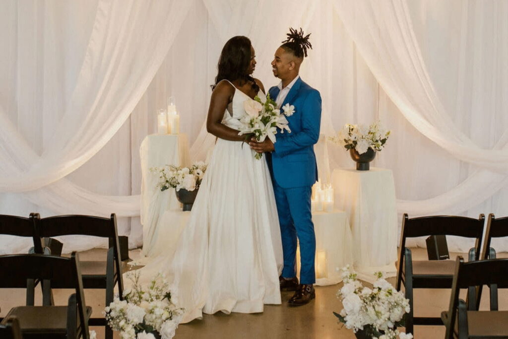 A bride and groom stand at a wedding altar draped in white linens and decorated with flowers and candles
