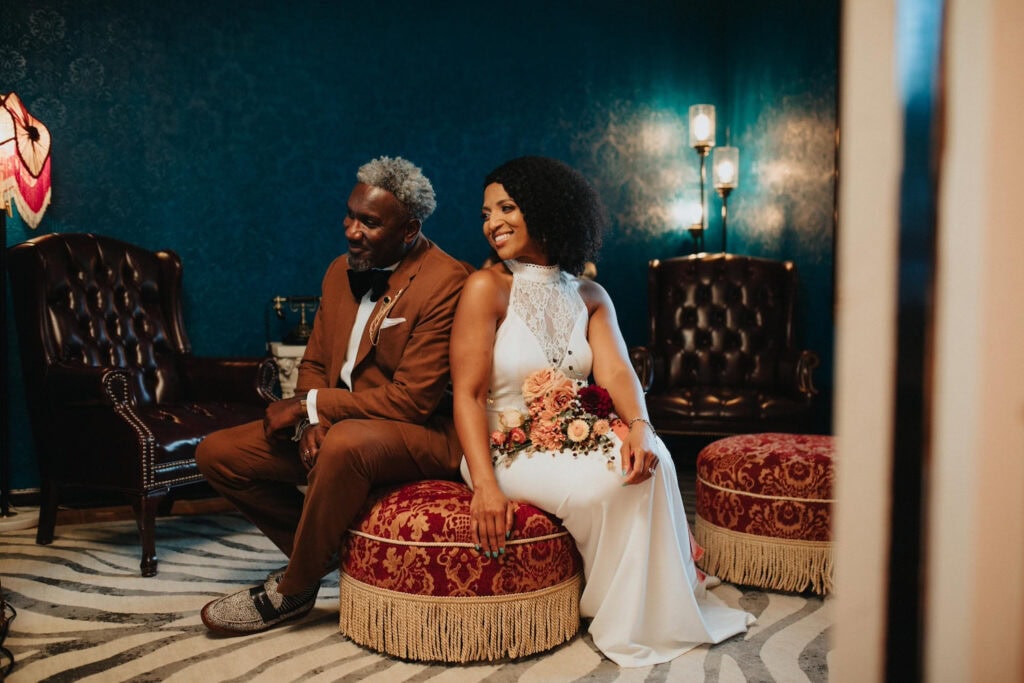 Newlyweds sit on a red and gold brocade ottoman in a dim room with aqua wallpaper and brown leather chairs