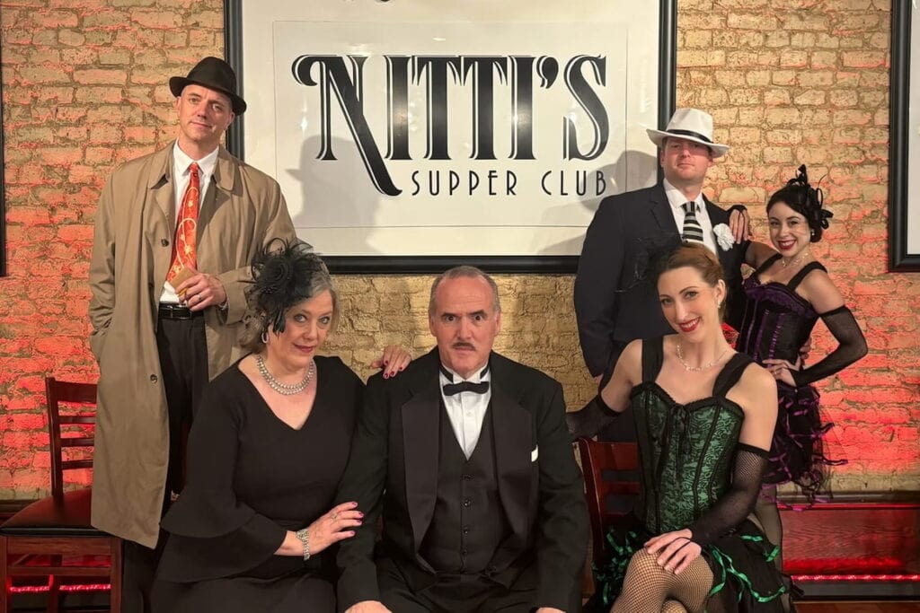 A group of people in Prohibition-era costumes stand by the Nitti's Supper Club sign