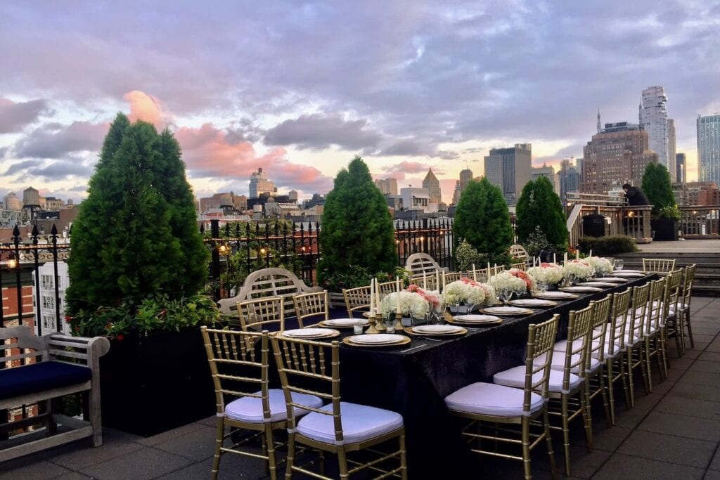 A long reception table is set on a rooftop in the city with tall buildings seen in the background