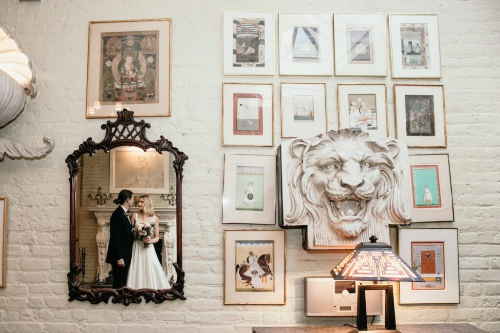 A painted brick wall with a bunch of framed prints surrounding a carving of a lion's head next to a mirror in which a bride and groom are seen in the reflection