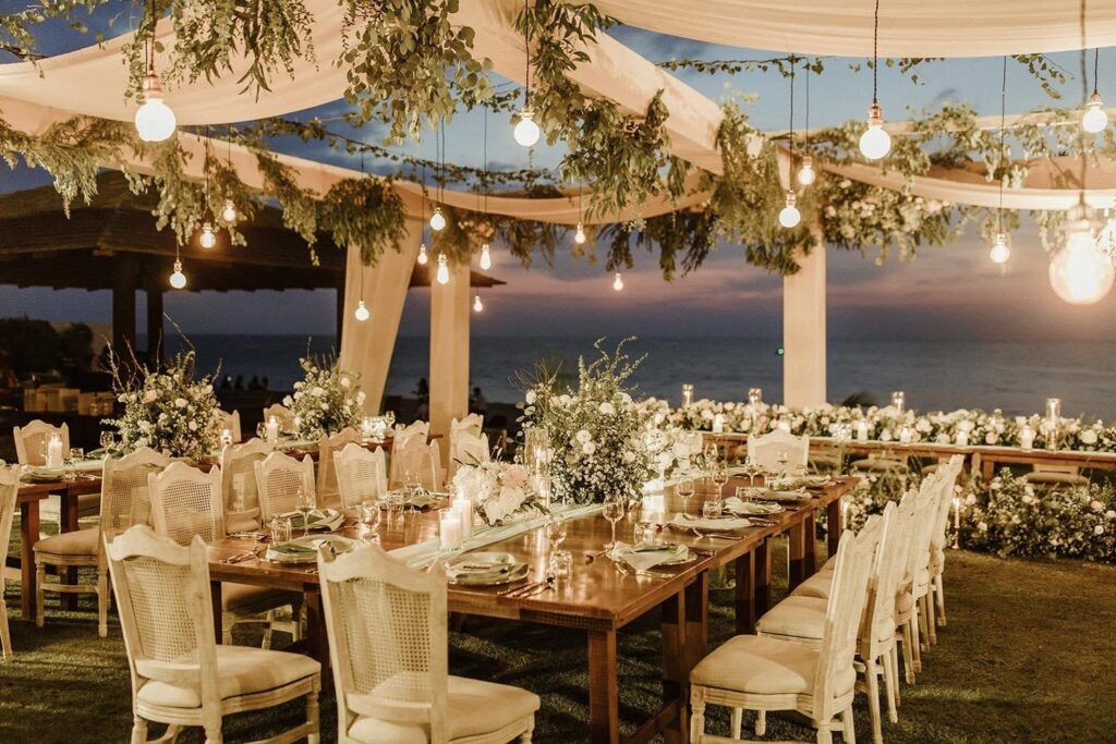 White wooden chairs flank large wooden tables at an outdoor reception by the sea under a canopy of greenery and bistro lights