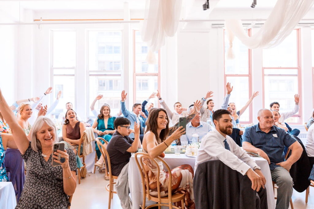 Wedding guests wave to the camera from their seats at a reception in a bright, light-filled room