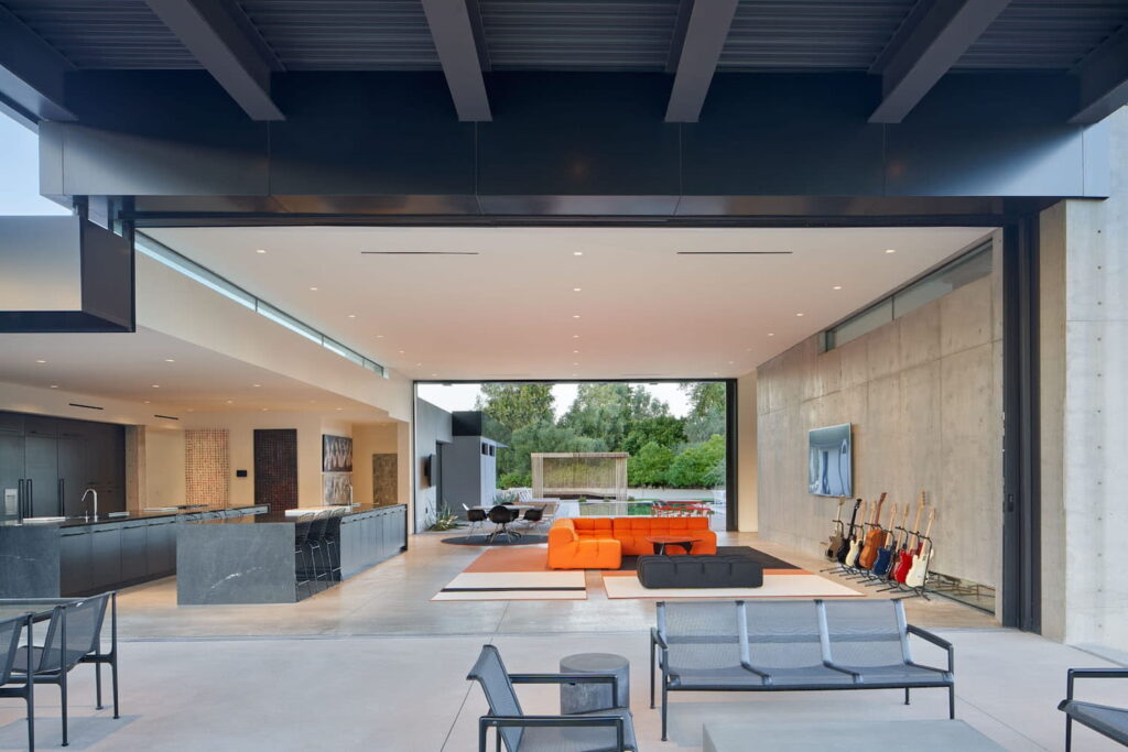 Open air living space in modern desert home with bright orange couch, huge doorways leading to patios and an all-black kitchen area