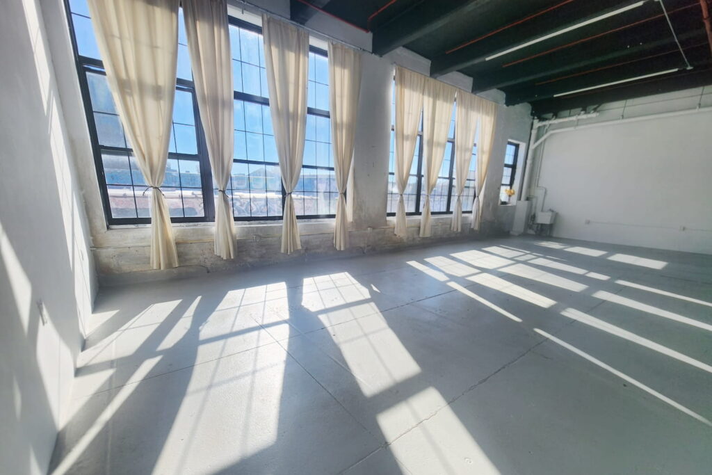 A large wall of windows draped in white curtains illuminates an all-white event space in Brooklyn