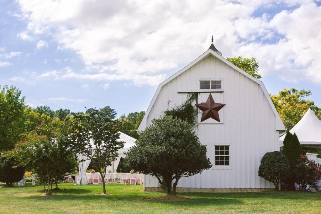 A white barn with a large metal star hanging on it sits next to a tented reception space decorated with white chairs and pink linens