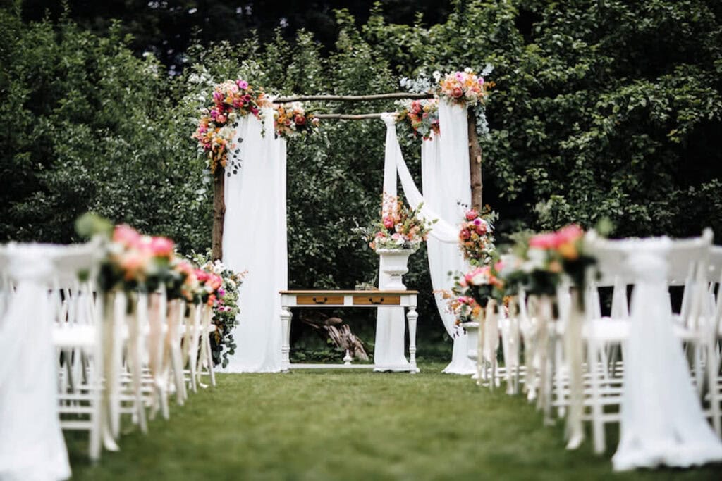 A wooden altar is decorated in white drapes and rich flowers at the front of two aisles of white chairs at an outdoor venue