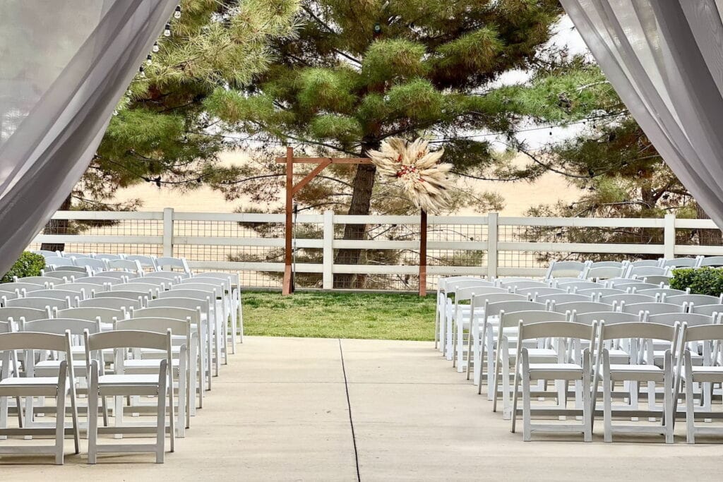 Two rows of white folding chairs flank an aisle leading towards a wooden altar decorated with a rustic, bohemian flower arrangement by a fence overlooking a field