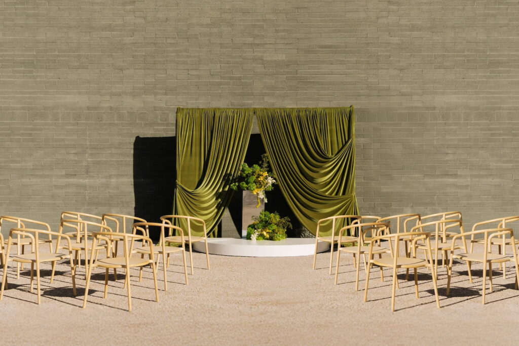 Light wooden chairs are lined up for a wedding ceremony at an outdoor venue by a large brick wall with a dramatic altar made of green velvet drapes and earthy flowers