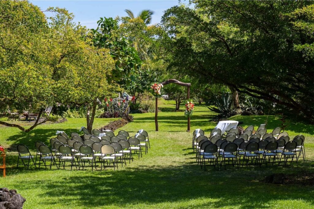 Chairs are set up in a grassy area by lush trees facing a wooden altar decorated with palms and bright flowers