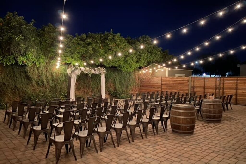 String lights twinkle above an outdoor ceremony set up with metal chairs, wine barrels and wooden altar