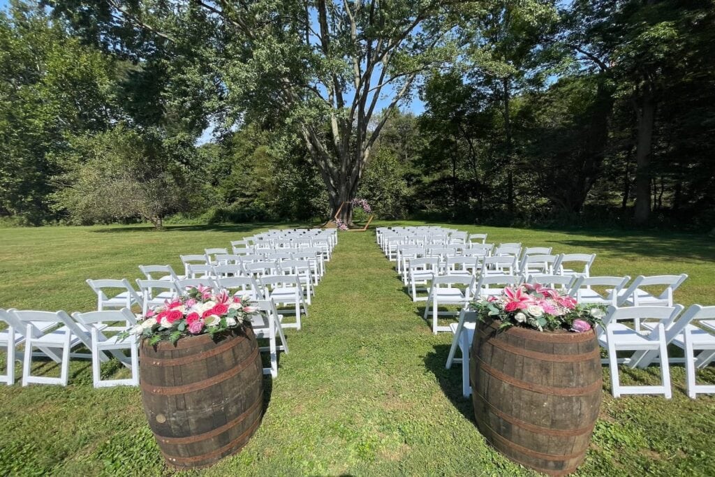 Large wine barrels accent an outdoor wedding ceremony space with white folding chairs and a quaint altar in the distance set up near a wooded area
